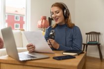 Delighted female radio host recording podcast while using mic and reading notes from paper at home — Stock Photo
