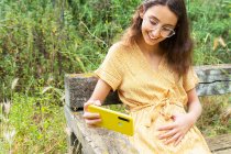 Улыбающаяся беременная женщина в платье трогает живот и делает автопортрет на мобильном телефоне, сидя летом на скамейке в сельской местности — стоковое фото