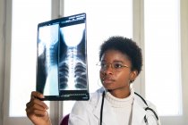 Giovane medico donna nera in piedi vicino alla finestra ed esaminando la scansione a raggi X mentre si lavora in clinica — Foto stock