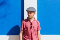 Продуманий молодий бородатий чоловік у стильному повсякденному одязі та кепці насолоджується літнім днем біля синьої стіни на міській вулиці — стокове фото
