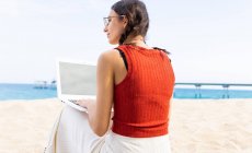 Back view of content female freelancer sitting on seashore and typing on laptop while working remotely on project in summer — Stock Photo