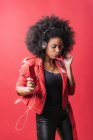 Delighted African American female listening to music in headphones and using mobile phone while dancing on red background in studio — Stock Photo