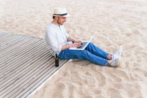 Vista lateral de cuerpo completo de un joven blogger con estilo y sombrero sentado con una botella de cerveza en la playa de arena y escribiendo en el portátil durante las vacaciones de verano en la orilla del mar - foto de stock