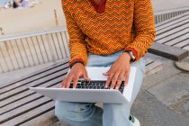 Обрезанный неузнаваемый положительный молодой этнический студент в стильном наряде, печатающий на клавиатуре ноутбука, сидя на скамейке у моря на городском пляже — стоковое фото