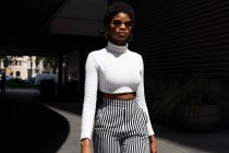Giovane femmina nera in abito elegante e occhiali da sole mentre cammina e guarda la fotocamera nella giornata di sole sulla strada della città — Foto stock