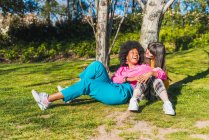 Pareja multiracial de mujeres homosexuales que se relajan en el prado en el parque mientras se abrazan y se divierten en un día soleado en el fin de semana de verano - foto de stock