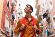 Bajo ángulo de confianza feliz joven étnico turista masculino con mochila y auriculares TWS mirando hacia otro lado mientras explora viejas calles estrechas de la ciudad de Barcelona - foto de stock