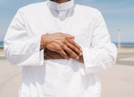 Crop Islamic male in traditional white clothes standing on rug and praying against blue sky on beach — Stock Photo