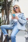 Trendige Frau im Jeans-Outfit sitzt auf der Straße und surft auf dem Handy in den sozialen Medien — Stockfoto