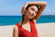 Fröhliche Frau in Sommerkleidung mit Zöpfen steht am sandigen Ufer mit ruhigem blauem Meer an einem sonnigen Tag und blickt in die Kamera — Stockfoto