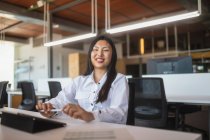 Positive asiatische Mitarbeiterin sitzt am Tisch im Arbeitsbereich und blickt in die Kamera — Stockfoto
