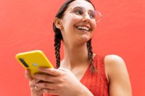 Par en bas joyeuse jeune femme en tresses coiffure navigation sur smartphone debout regardant loin sur fond rouge dans la rue — Photo de stock
