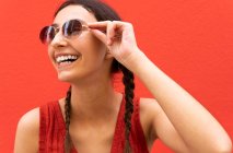 Délicieuse jeune femme avec des nattes portant des lunettes de soleil à la mode sur fond rouge dans la rue de la ville — Photo de stock