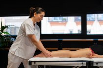 Vista laterale della massaggiatrice felice in accappatoio bianco massaggiare polpaccio di coltura paziente femminile durante la sessione di fisioterapia in clinica — Foto stock