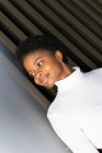 Élégante jeune femme noire en pull de culture regardant loin tout en se tenant près du mur de construction rayé en ville — Photo de stock