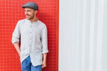 Bonito jovem barbudo cara na moda camisa casual e jeans e boné de pé perto da parede vermelha e branca e olhando para longe com sorriso — Fotografia de Stock