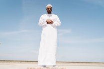 Повний хлопчик Ісламський чоловік в традиційному білому одязі стоїть на килимі і молиться проти блакитного неба на пляжі. — стокове фото