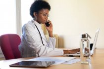 Competent young African American female medical practitioner answering phone call and using laptop while consulting patients remotely from office — Stock Photo