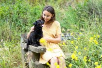 Mulher alegre com cachorro fofo preto sentado no banco de madeira no campo — Fotografia de Stock