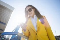 Усміхнена азіатська бізнес-леді з жовтим пальто і смартфон, що йде на вулиці з будівлею на задньому плані — стокове фото