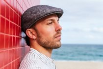Vista lateral de un hombre barbudo joven reflexivo con elegante camisa a rayas y sombrero de pie mirando hacia otro lado cerca de la pared roja y disfrutando del soleado día de verano en la calle - foto de stock