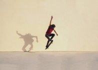Hombre skateboarder con el pelo ondulado realizar truco en el monopatín mientras salta sobre la pasarela y mirando hacia abajo en el día soleado - foto de stock