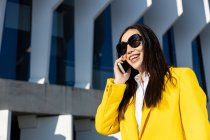 Lächelnde asiatische Geschäftsfrau mit gelbem Mantel und Smartphone, die auf der Straße läuft, mit einem Gebäude im Hintergrund — Stockfoto