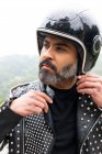 Auto assegurada adulto barbudo masculino motociclista na jaqueta de couro elegante ajustando capacete protetor e olhando para longe na natureza — Fotografia de Stock