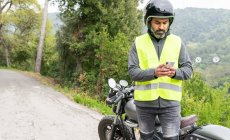 Вважний дорослий іспаномовний велосипедист в захисному шоломі і жилети повідомлення на мобільному телефоні стоячи біля розбитого мотоцикла біля буйних зелених лісів. — стокове фото
