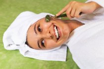 Top view joven hembra feliz con toalla en la cabeza sonriendo y masajeando la cara con rodillo de jade durante la rutina de cuidado de la piel en casa - foto de stock