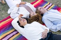 High angle of diverse women and man with curly hair lying face to face on colorful plaid in park looking up — Stock Photo