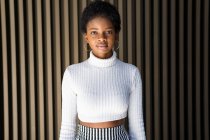Stilvolle junge schwarze Frau im Crop-Pullover blickt in die Kamera, während sie in der Nähe einer gestreiften Hauswand in der Stadt steht — Stockfoto