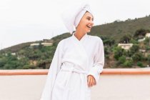 Ottimista giovane donna in accappatoio e asciugamano sorridente e distogliendo lo sguardo mentre si rilassa sul balcone durante la routine di cura della pelle nel fine settimana — Foto stock