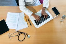 Из выше урожая афроамериканка врач печатает отчет на ноутбуке во время работы за столом в современном офисе клиники — стоковое фото
