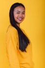 Vista lateral da mulher asiática encantada em pé no fundo amarelo no estúdio enquanto olha para a câmera — Fotografia de Stock