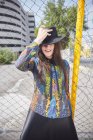 Contenido femenino en ropa de moda de pie cerca de valla de malla en la ciudad en el día soleado y cubriendo los ojos con sombrero - foto de stock