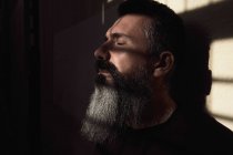 Вид сбоку мечтательного взрослого бородатого мужчины с закрытыми глазами в темной комнате с солнечным светом — стоковое фото