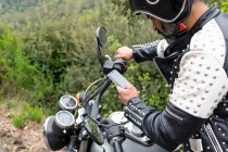 Vista laterale di motociclista maschio in giacca di pelle alla moda con rivetti e casco protettivo telefono di navigazione mentre seduto su moto parcheggiata vicino a lussureggiante foresta verde — Foto stock