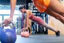 Jóvenes atletas femeninas decididas en ropa deportiva de pie en postura de tablón durante el entrenamiento funcional con pesas en el suelo en el gimnasio - foto de stock