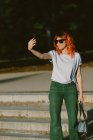 Trendy female with ginger hair and in sunglasses taking self shot on mobile phone on sunny day in street — Stock Photo
