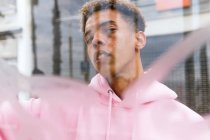 Durch das Glas eines ernsthaften jungen ethnischen Hipsters mit Afro-Frisur, gekleidet in rosa Kapuzenpullover am Fenster und in die Kamera schauend — Stockfoto