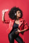 Encantada mujer afroamericana escuchando música en auriculares y usando el teléfono móvil mientras baila sobre fondo rojo en el estudio - foto de stock