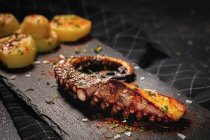 Du dessus du tentacule de poulpe frit et des morceaux de pomme de terre servis avec des épices sur un plateau noir sur la table — Photo de stock