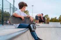 Вид збоку хлопчика сидить зі скейтбордом на рампі в скейт-парку і дивиться в сторону — стокове фото