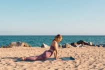 Side view of female doing yoga in Bhujangasana and stretching body on sandy beach on sunny day — Stock Photo