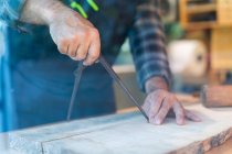 Crop unrecognizable male woodworker using professional compass or divider while marking wooden plank at workbench in carpentry workshop — Stock Photo