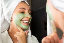Mulher feliz com toalha na cabeça sorrindo e espalhando máscara verde no rosto enquanto olha para o espelho no banheiro em casa — Fotografia de Stock