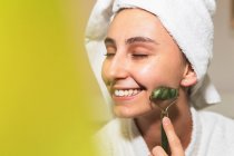 Happy young female with towel on head smiling and massaging face with jade roller during skin care routine at home — Stock Photo