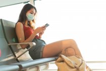 Turista con máscara protectora sentada en la sala de salida del aeropuerto y esperando el vuelo durante la epidemia de coronavirus mientras usa el teléfono inteligente - foto de stock