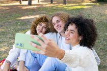 Grupo de mujeres multirraciales jóvenes felices y hombre con el pelo rizado sentado en la hierba verde en el parque mientras toma selfie con el teléfono móvil - foto de stock
